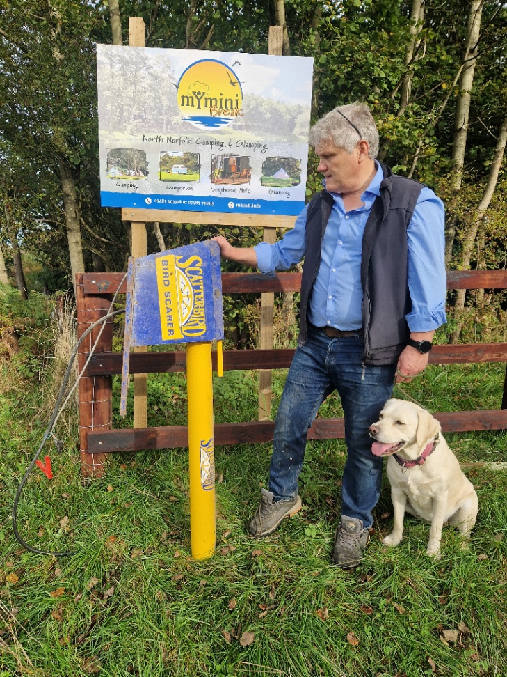Nigel Marsh standing next to a yellow Scatterbird gas-powered bird scarer mounted on a post at Dam Hill near Holt, with his black Labrador sitting beside him and the mYminiBreak sign visible behind