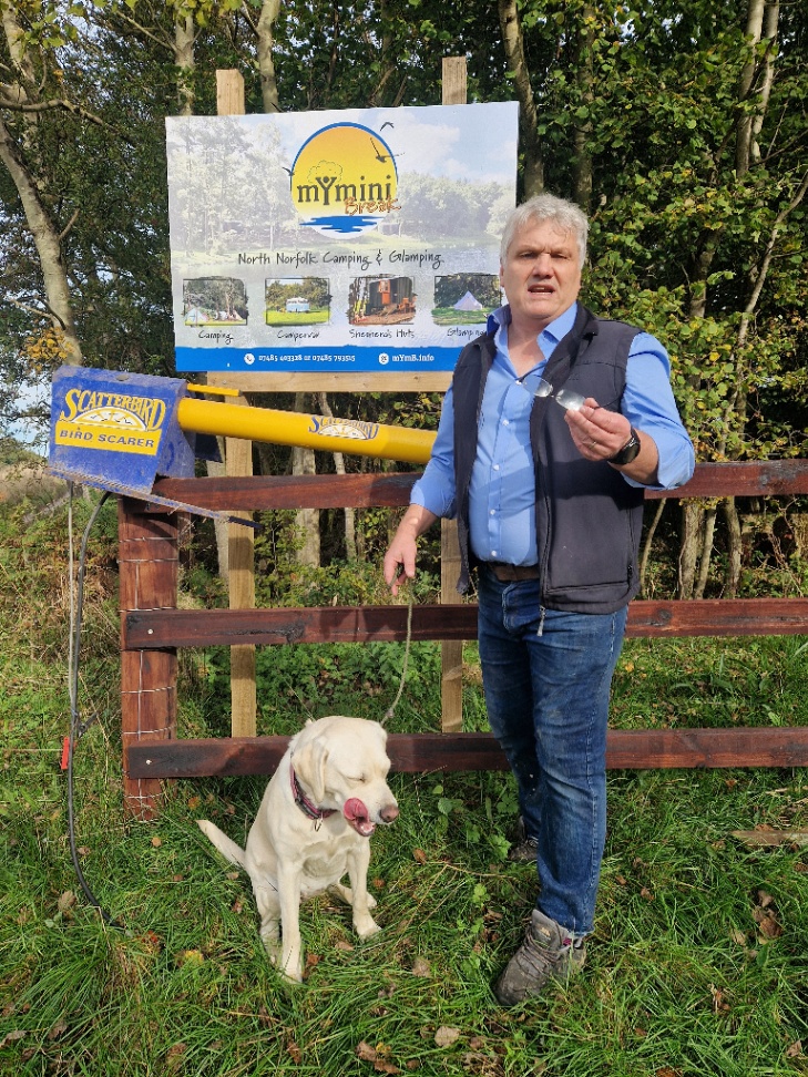 Nigel Marsh holding up his business card next to a Scatterbird bird scarer post at Dam Hill near Holt, with his Labrador on a lead beside him