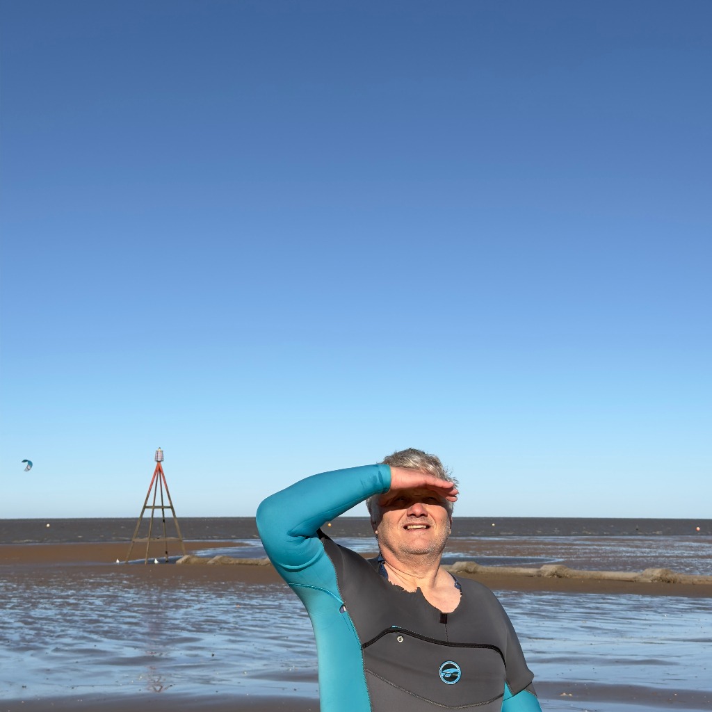 Nigel Marsh in grey and teal wetsuit on Hunstanton beach flats North Norfolk UK smiling hand to brow clear sky over The Wash